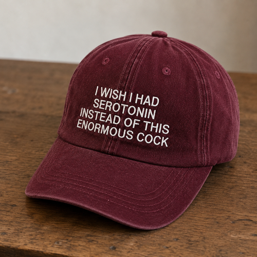 A maroon cotton baseball cap with white embroidered text reading 'I WISH I HAD SEROTONIN INSTEAD OF THIS ENORMOUS COCK' resting on a wooden surface.