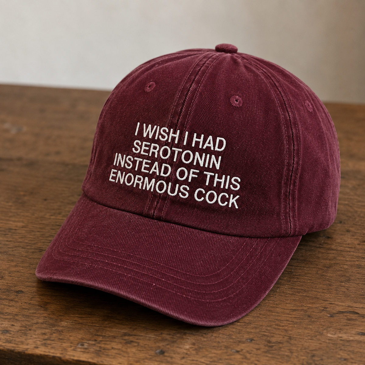 A maroon cotton baseball cap with white embroidered text reading 'I WISH I HAD SEROTONIN INSTEAD OF THIS ENORMOUS COCK' resting on a wooden surface.