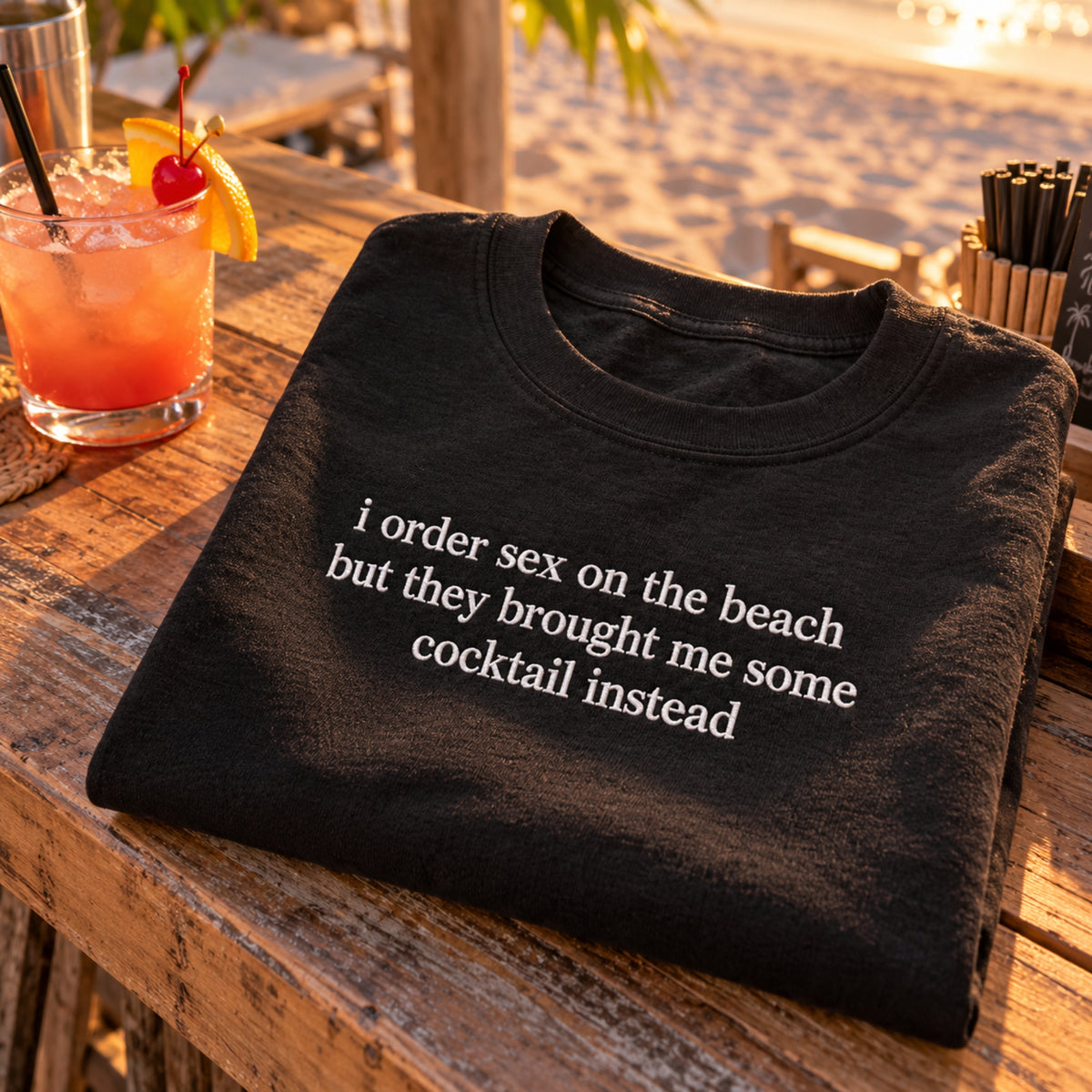 A folded black t-shirt with white text reading 'i order sex on the beach but they brought me some cocktail instead' is displayed on a wooden beachside table next to a cocktail glass.