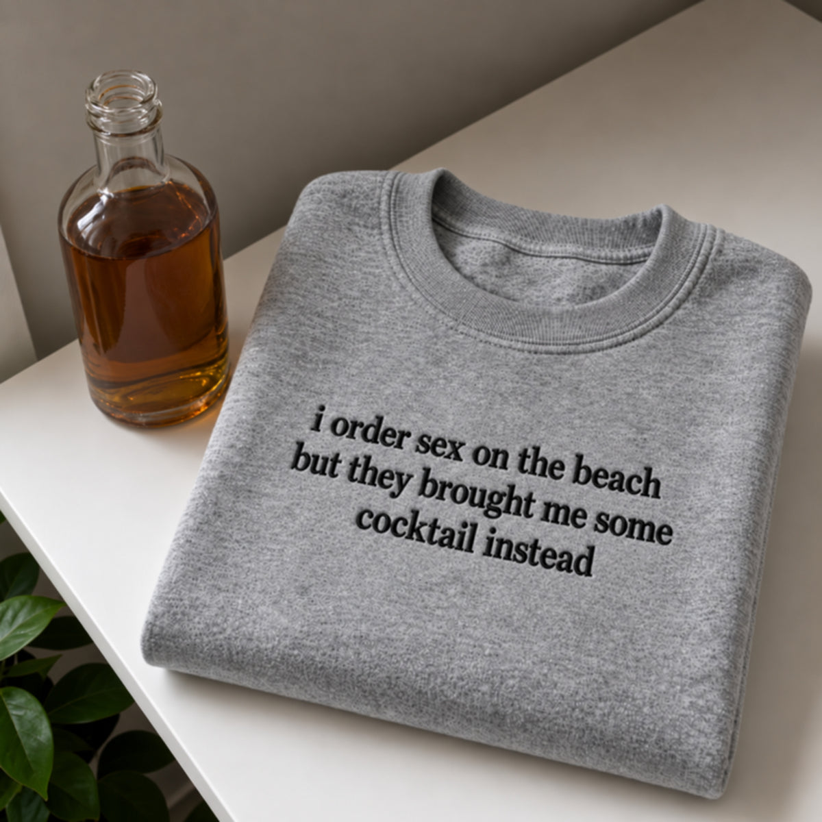 A folded heather gray crewneck sweatshirt with black text reading 'i order sex on the beach but they brought me some cocktail instead' sits on a white tabletop next to a glass bottle of amber liquid, with a small green plant leaf visible at the lower left.
