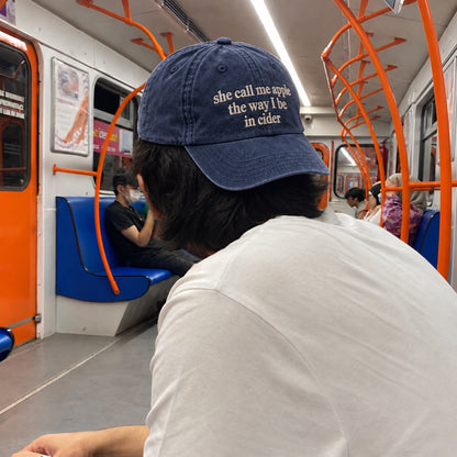 A person sitting inside a subway train wearing a navy blue baseball cap with the phrase 'she call me apple the way I be in cider' in beige text. The photo is taken from behind the person, showing their white shirt and the interior of the train with orange handrails and blue seats.