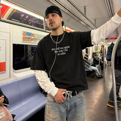 A man standing on a subway train wearing a black graphic t-shirt with the text 'she call me apple the way the in cider' in white, layered over a white long-sleeve shirt, light wash jeans, and a black beanie.