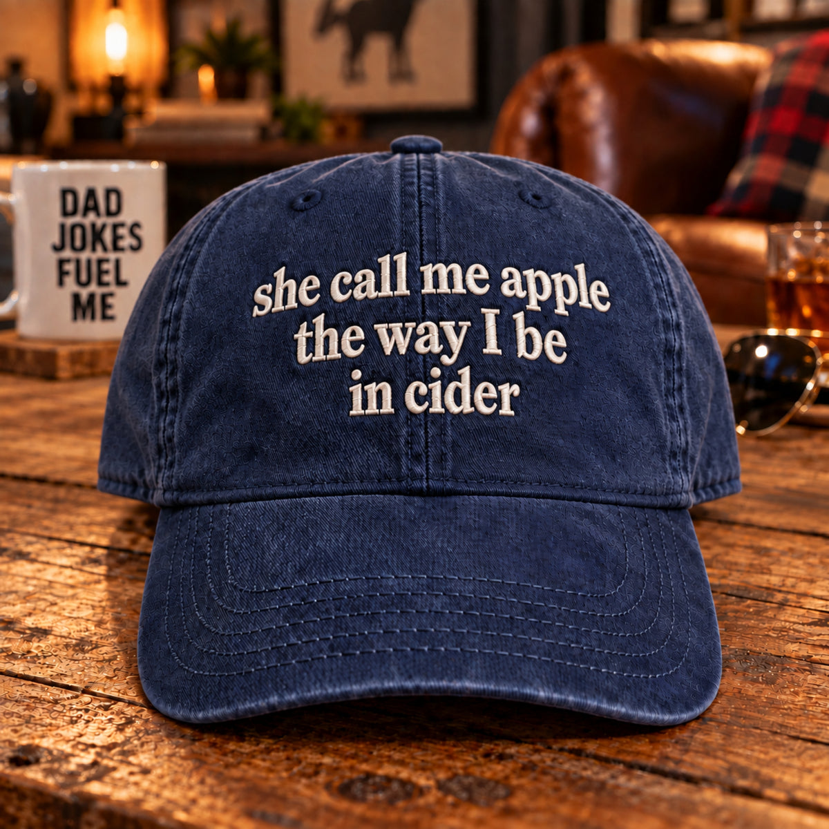 A navy blue washed baseball cap with white embroidered text reading 'she call me apple the way I be in cider' sits on a rustic wooden table in a cozy indoor setting with a mug, sunglasses, and leather furniture in the background.