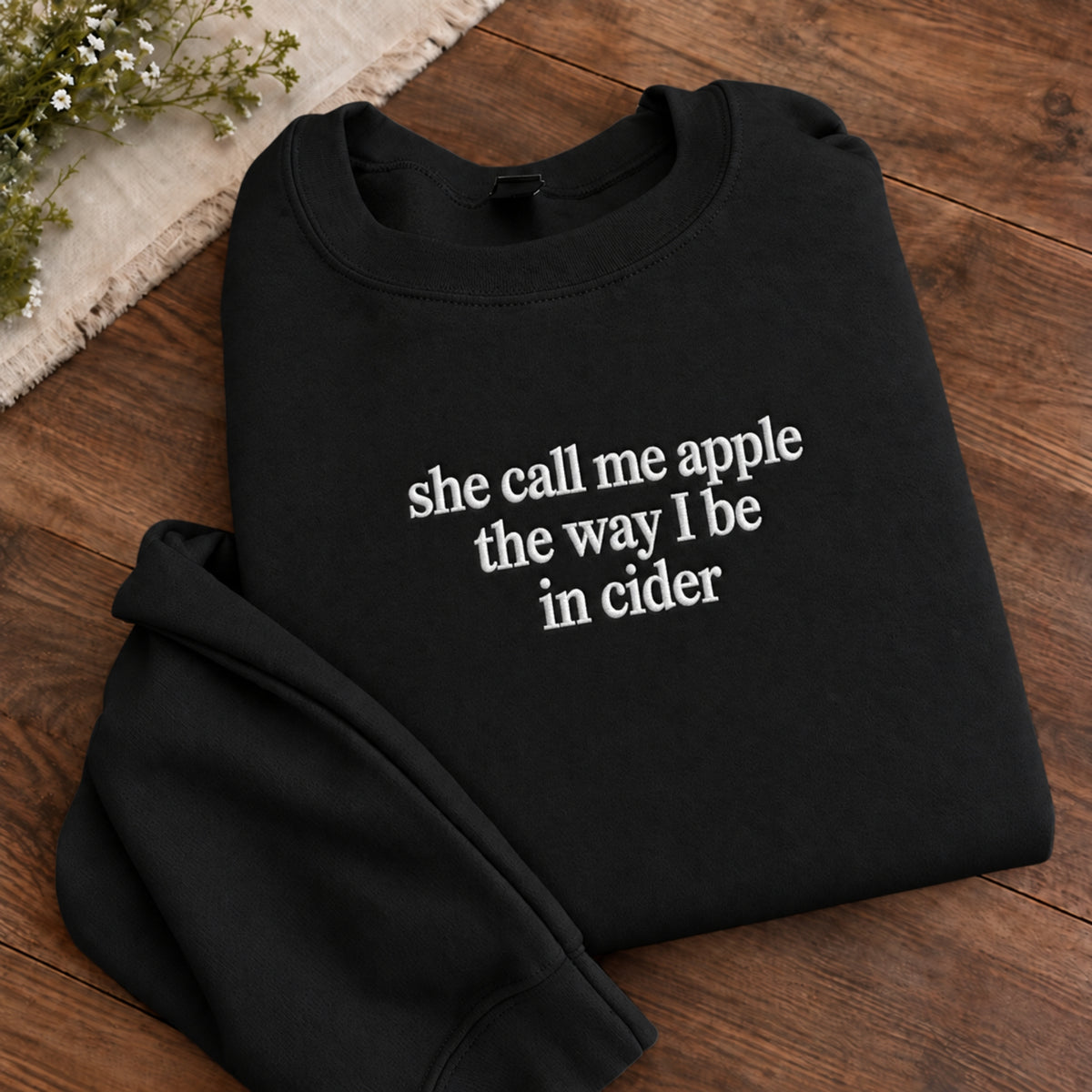 A folded black crewneck sweatshirt with white text reading 'she call me apple the way I be in cider' on a wooden surface, styled with a small sprig of flowers in the corner.