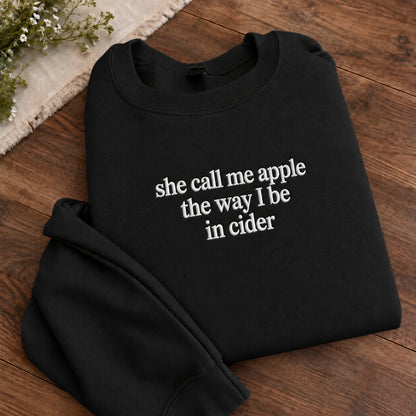 A folded black crewneck sweatshirt with white text reading 'she call me apple the way I be in cider' on a wooden surface, styled with a small sprig of flowers in the corner.