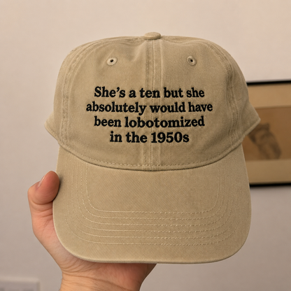 A hand holding a beige baseball cap with black embroidered text that reads, 'She's a ten but she absolutely would have been lobotomized in the 1950s'.