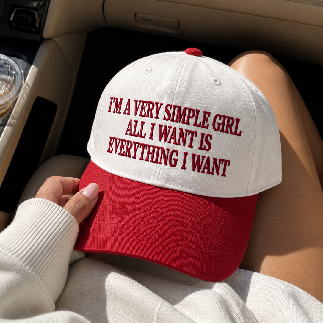A white and red baseball cap with the text 'I'M A VERY SIMPLE GIRL ALL I WANT IS EVERYTHING I WANT' in red embroidery, held by a person sitting inside a car.