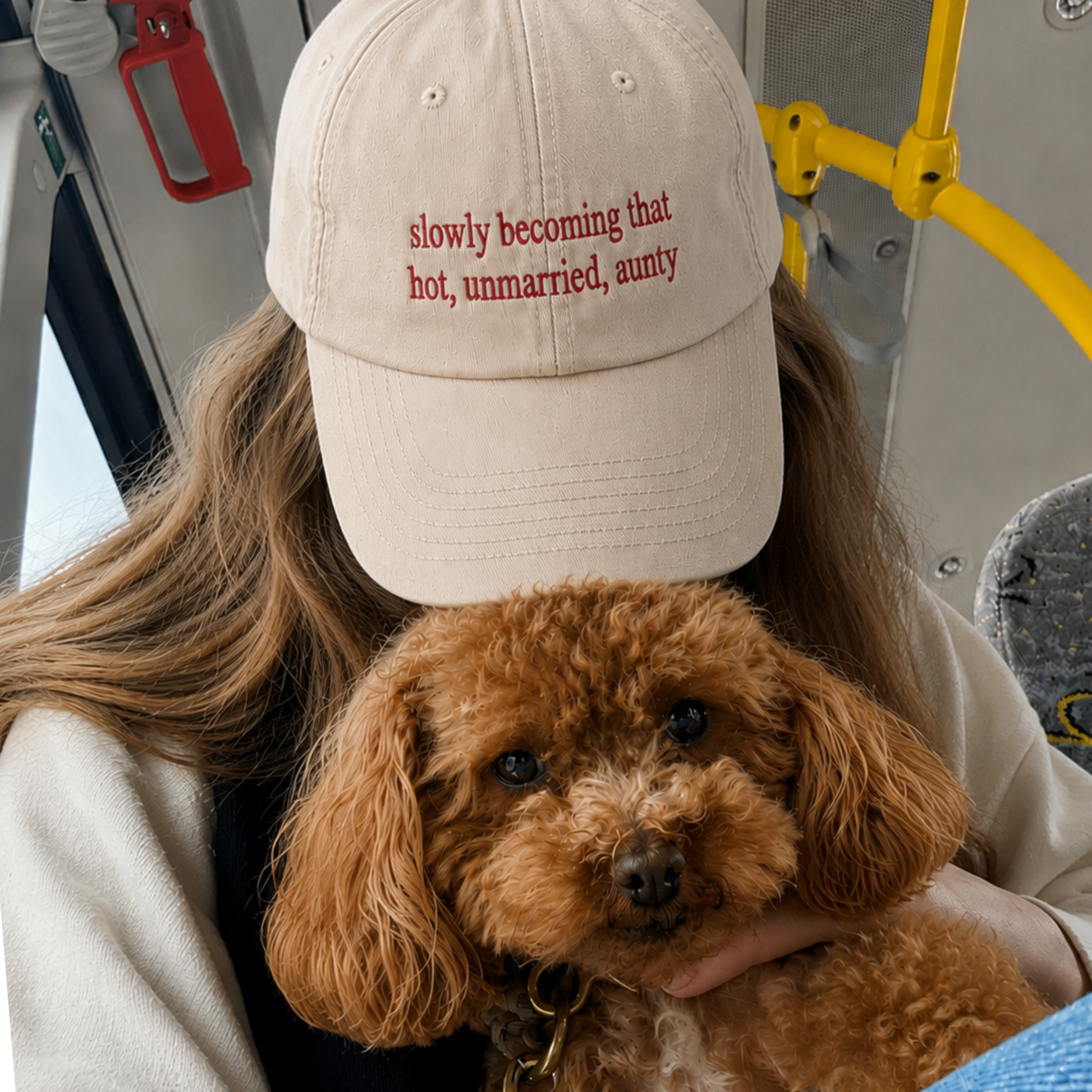A person wearing a beige baseball cap with red embroidered text 'slowly becoming that hot, unmarried, aunty' holds a fluffy brown poodle on a bus. The person is wearing a light beige jacket, and the background shows yellow bus handrails and gray interior details.