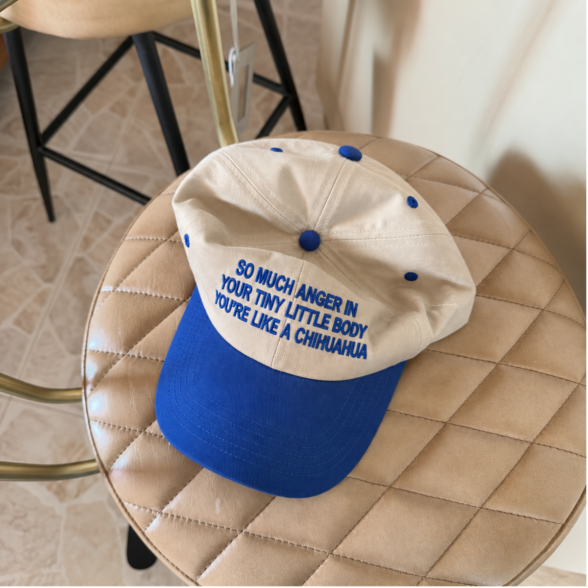 A beige and royal blue baseball cap with blue embroidered text reading 'SO MUCH ANGER IN YOUR TINY LITTLE BODY YOU'RE LIKE A CHIHUAHUA', resting on a beige quilted stool.
