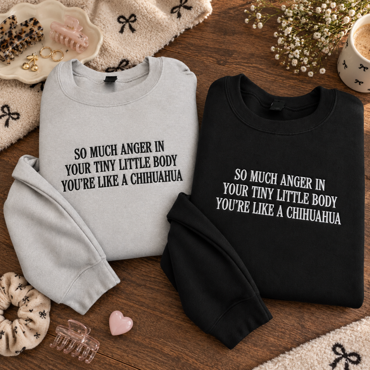 Two folded sweatshirts with the same slogan in different colors are arranged on a wooden surface with cozy props. The left sweatshirt is light gray with black text reading 'SO MUCH ANGER IN YOUR TINY LITTLE BODY YOU'RE LIKE A CHIHUAHUA.