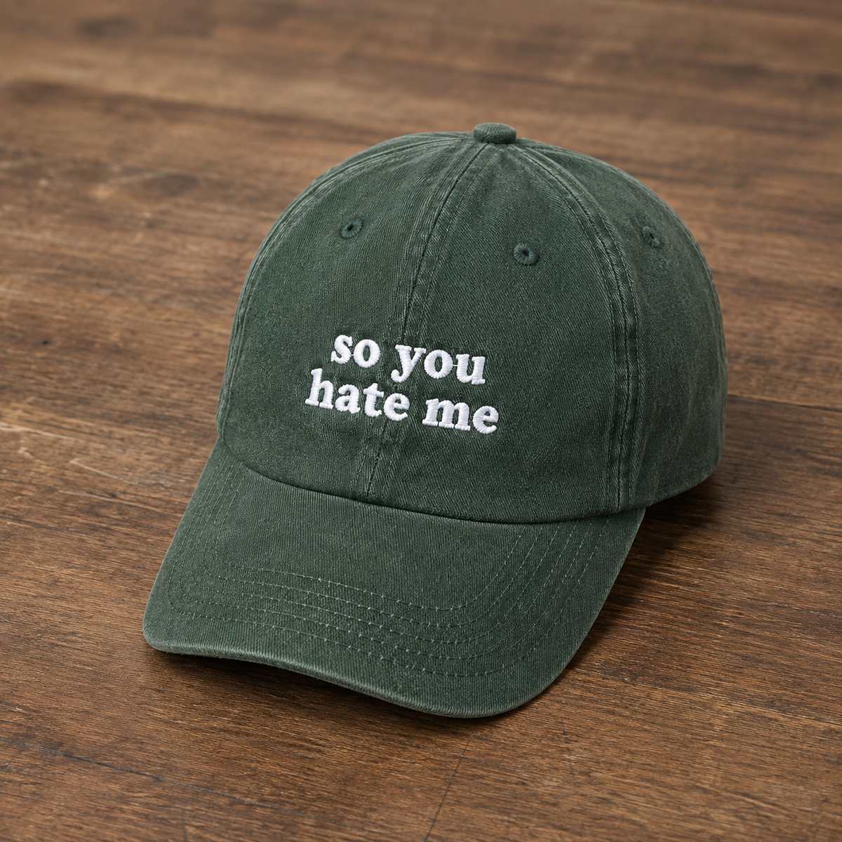 A dark green baseball cap with white embroidered text reading 'so you hate me' on the front, displayed on a brown wooden surface.