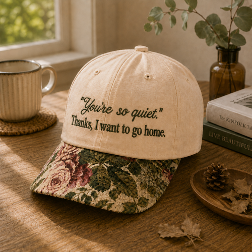 Beige embroidered baseball cap with black text reading "You're so quiet. Thanks, I want to go home." featuring a vintage floral tapestry brim, displayed on a wooden surface next to a cozy mug and books.