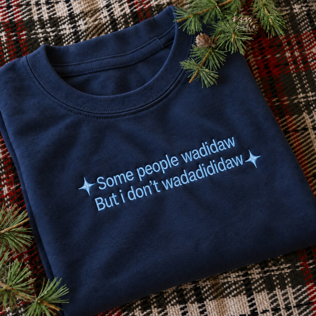 A navy blue folded t-shirt with light blue embroidered text reading 'Some people wadidaw But i don't wadadididaw' flanked by small star sparkles, styled on a red and black plaid blanket with pine branches.