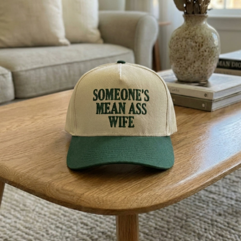 A two-tone baseball cap with a cream-colored crown and a forest green visor, featuring the text 'SOMEONE'S MEAN ASS WIFE' embroidered in green on the front. The cap is styled on a wooden table in a cozy living room setting.