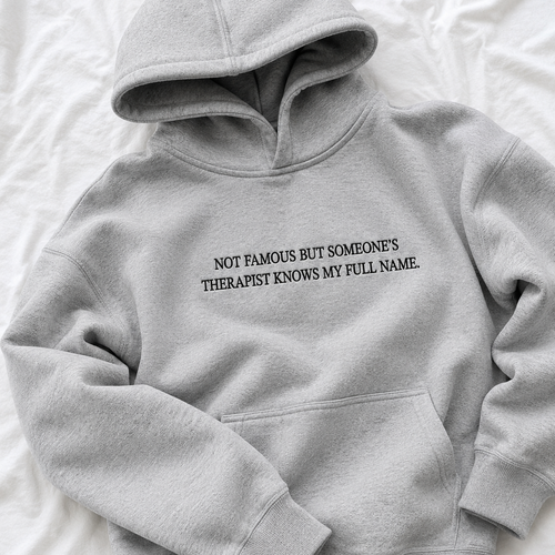 A light gray pullover hoodie with black printed text on the chest reading 'NOT FAMOUS BUT SOMEONE'S THERAPIST KNOWS MY FULL NAME.' laid flat on a white fabric background.