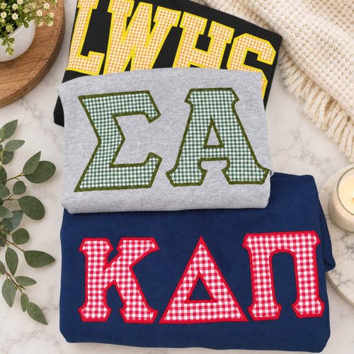 Three folded sweatshirts stacked on a marble surface with decorative props. The top sweatshirt is black with yellow gingham letters reading "LWHS," the middle is light gray with green gingham letters reading "ΣΑ," and the bottom is navy blue with red gingham letters reading "ΚΑΠ.