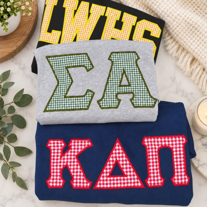 Three folded sweatshirts stacked on a marble surface with decorative props. The top sweatshirt is black with yellow gingham letters reading "LWHS," the middle is light gray with green gingham letters reading "ΣΑ," and the bottom is navy blue with red gingham letters reading "ΚΑΠ.