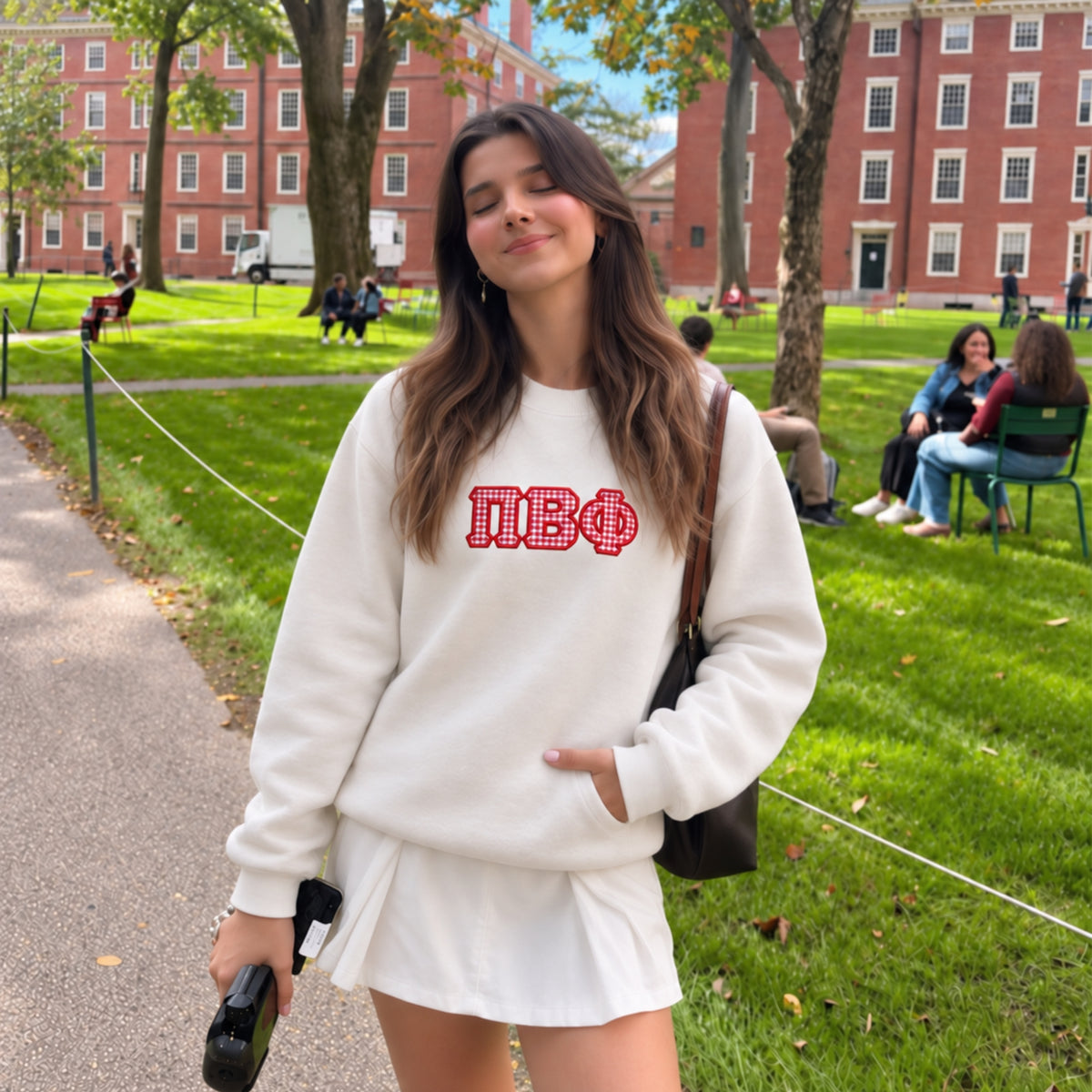 A woman stands on a campus walkway wearing a white sweatshirt and matching white skirt with a red Greek letter graphic on the chest. The background shows a green lawn, trees, and red brick buildings.