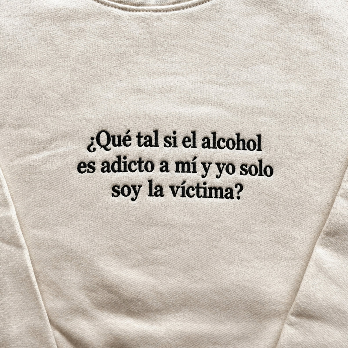 Cream sweatshirt with black embroidered Spanish quote “¿Qué tal si el alcohol es adicto a mí y yo solo soy la víctima?” centered on the chest in a bold serif font.