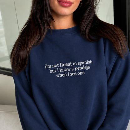 A woman wearing a navy blue crewneck sweatshirt with white text that reads 'i'm not fluent in spanish but i know a pendeja when i see one'.