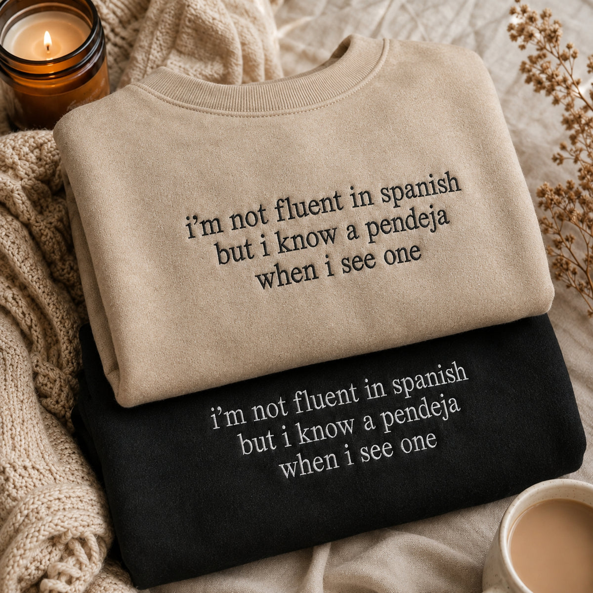 Two folded crewneck sweatshirts with Spanish text are displayed in a cozy flat-lay setting. The top sweatshirt is beige with black text, and the bottom is black with white text.