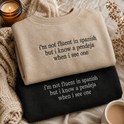 Two folded crewneck sweatshirts with Spanish text are displayed in a cozy flat-lay setting. The top sweatshirt is beige with black text, and the bottom is black with white text.