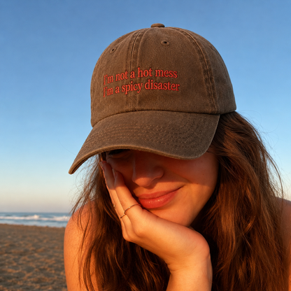 A woman on a beach wearing a brown baseball cap with red embroidered text that reads 'I'm not a hot mess I'm a spicy disaster'.