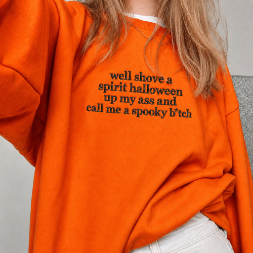 A close-up of a person wearing an oversized bright orange sweatshirt with black text on the chest that reads, 'well shove a spirit halloween up my ass and call me a spooky b*tch'.
