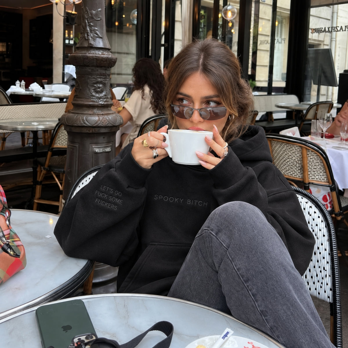 A woman wearing a black graphic hoodie and gray jeans sits at an outdoor cafe table, holding a white coffee mug. The hoodie features the text 'SPOOKY BITCH' and 'LET'S DO ENJOYING THINGS' on the front.