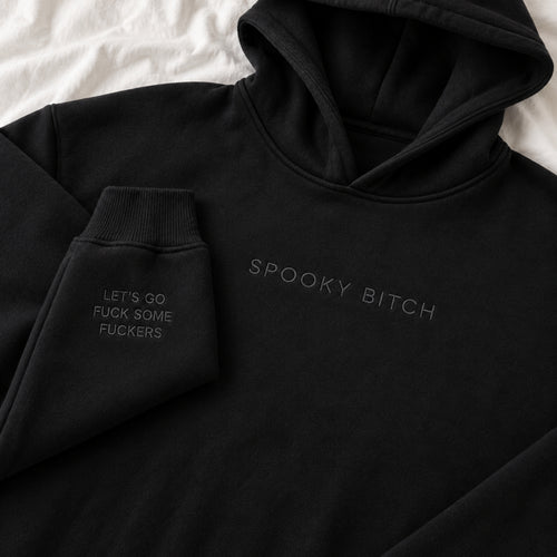 A black pullover hoodie laid flat on a white fabric background, with tonal embroidered text reading 'SPOOKY BITCH' across the chest and additional text on the left cuff reading 'LET'S GO FUCK SOME FUCKERS'.