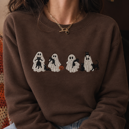 A person wearing a brown crewneck sweatshirt with a graphic of four white ghosts carrying black cats, with a small orange pumpkin, on a dark background.