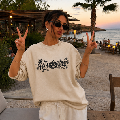 A woman posing at a beachside cafe wearing an oversized cream-colored sweatshirt with a black 'Halloween' graphic print featuring a jack-o'-lantern. She is making peace signs with both hands, wearing sunglasses, and the background shows a sunset over the ocean with palm trees.