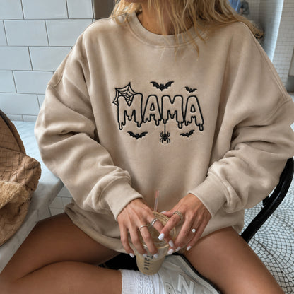 A woman wearing a beige crewneck sweatshirt with a black 'MAMA' graphic and Halloween-themed details, holding an iced coffee while sitting in a chair.