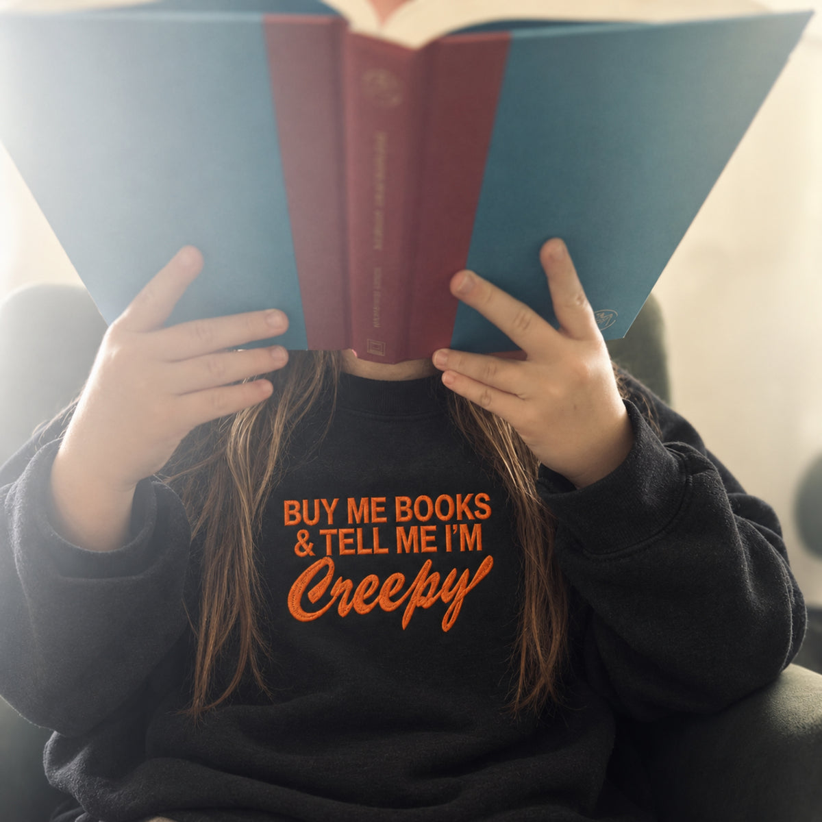 A child wearing a black sweatshirt with orange text that reads 'BUY ME BOOKS & TELL ME I'M Creepy' holds an open book in front of their face. The book has a blue cover on the left and a burgundy-red center spine/cover on the right.