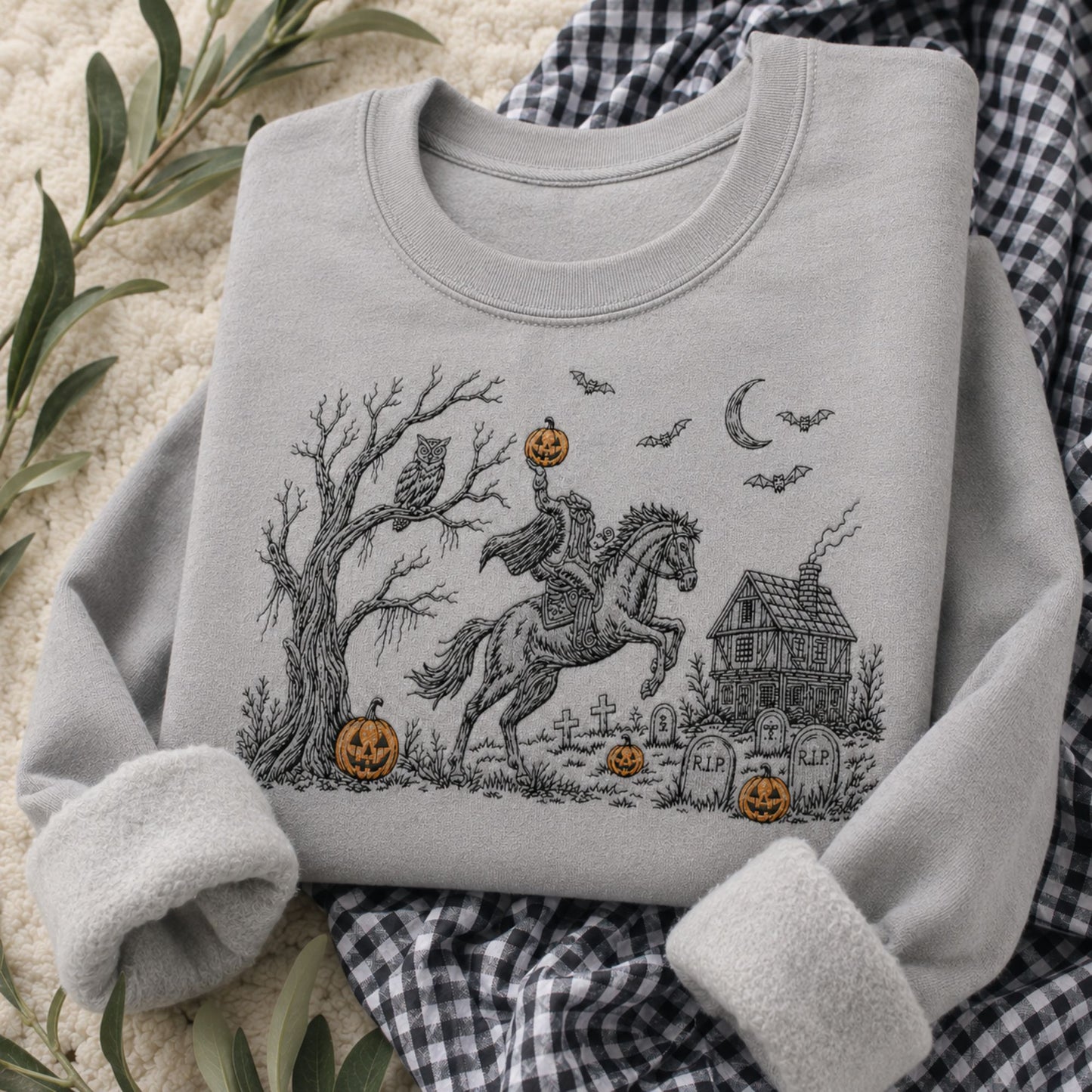 A light gray long-sleeve sweatshirt with a Halloween-themed graphic print, styled in a flat-lay arrangement with a black and white gingham blanket, green leaves, and a cream textured background. The graphic shows a winged horse, pumpkins, a haunted house, bats, and an owl.