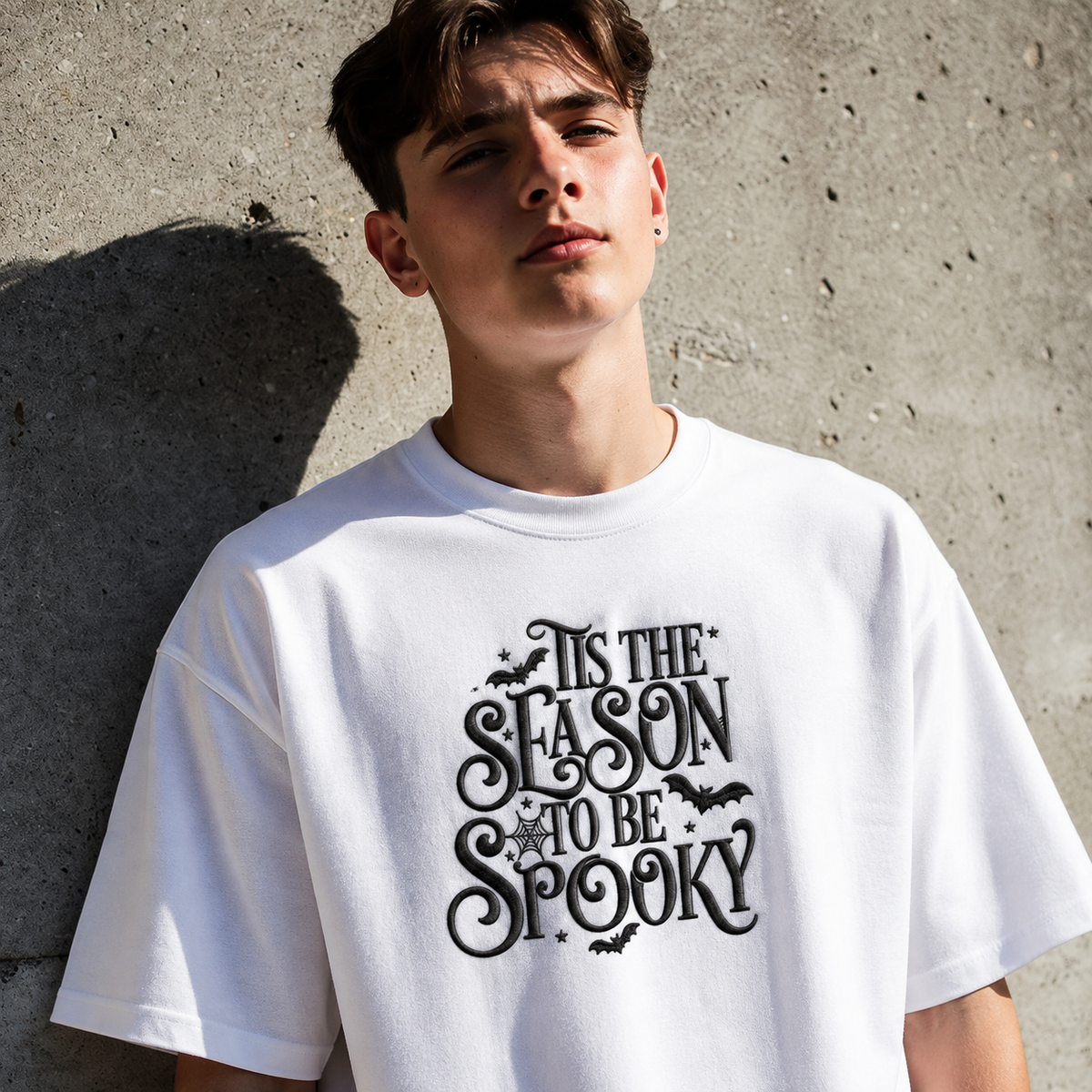 A young man wearing a white t-shirt with a black graphic that reads 'IT IS THE SEASON TO BE SPOOKY' with bats and stars, leaning against a concrete wall.