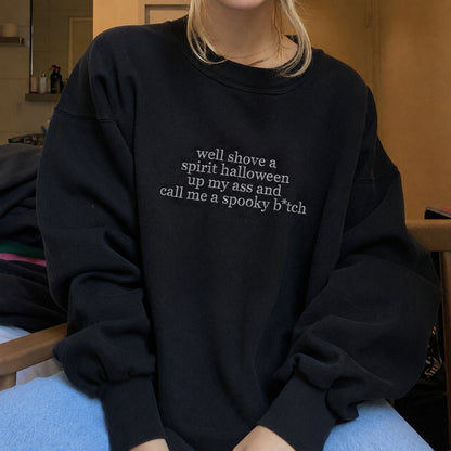 A person wearing an oversized black sweatshirt with white text on the chest that reads, 'well shove a spirit halloween up my ass and call me a spooky b*tch'.