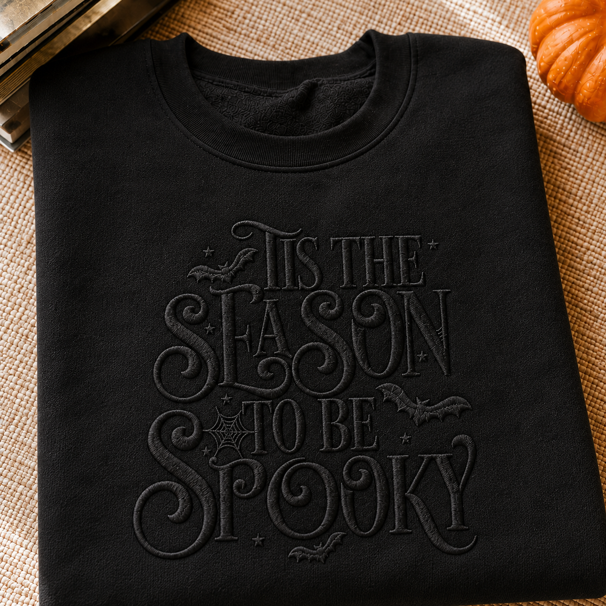 A folded black crewneck sweatshirt with tonal black embroidery reading 'It's the season to be spooky' with bats and spiderwebs, displayed on a burlap surface with a small pumpkin prop.