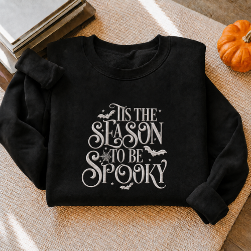 A black crewneck sweatshirt with a white graphic reading 'IT'S THE SEASON TO BE SPOOKY' with bats and stars, styled on a woven beige blanket with books and a small pumpkin nearby.