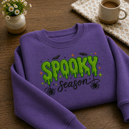 A purple crewneck sweatshirt with a green 'Spooky Season' graphic printed on the front, styled on a wooden tabletop with a small potted daisy, a mug of coffee, and a patterned napkin.