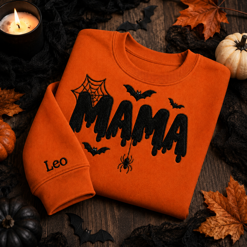 Bright orange Halloween crewneck sweatshirt with black embroidered "MAMA" dripping lettering, cobwebs, bats, a dangling spider, and custom name embroidery on the sleeve, styled on a flat lay with autumn leaves, pumpkins, and a lit candle.
