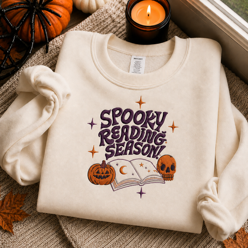 A cream-colored sweatshirt with a 'Spooky Reading Season!' graphic featuring a purple book, pumpkin, and skull, styled in a cozy autumn setting with a candle, small pumpkins, and leaves.