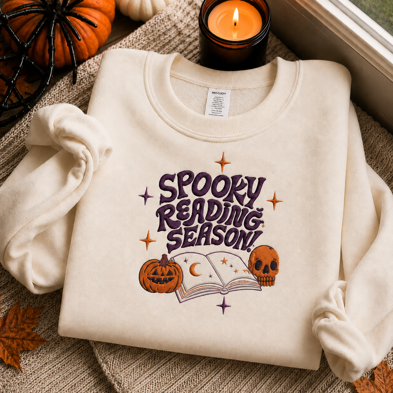 A cream-colored sweatshirt with a 'Spooky Reading Season!' graphic featuring a purple book, pumpkin, and skull, styled in a cozy autumn setting with a candle, small pumpkins, and leaves.