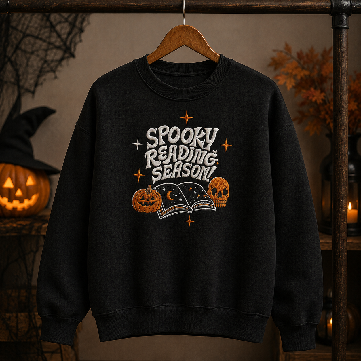A black crewneck sweatshirt hanging on a wooden hanger, featuring a 'Spooky Reading Season' graphic with a pumpkin, skull, open book, and stars. The background is a dimly lit, cozy Halloween-themed setting with a jack-o'-lantern and warm ambient lights.