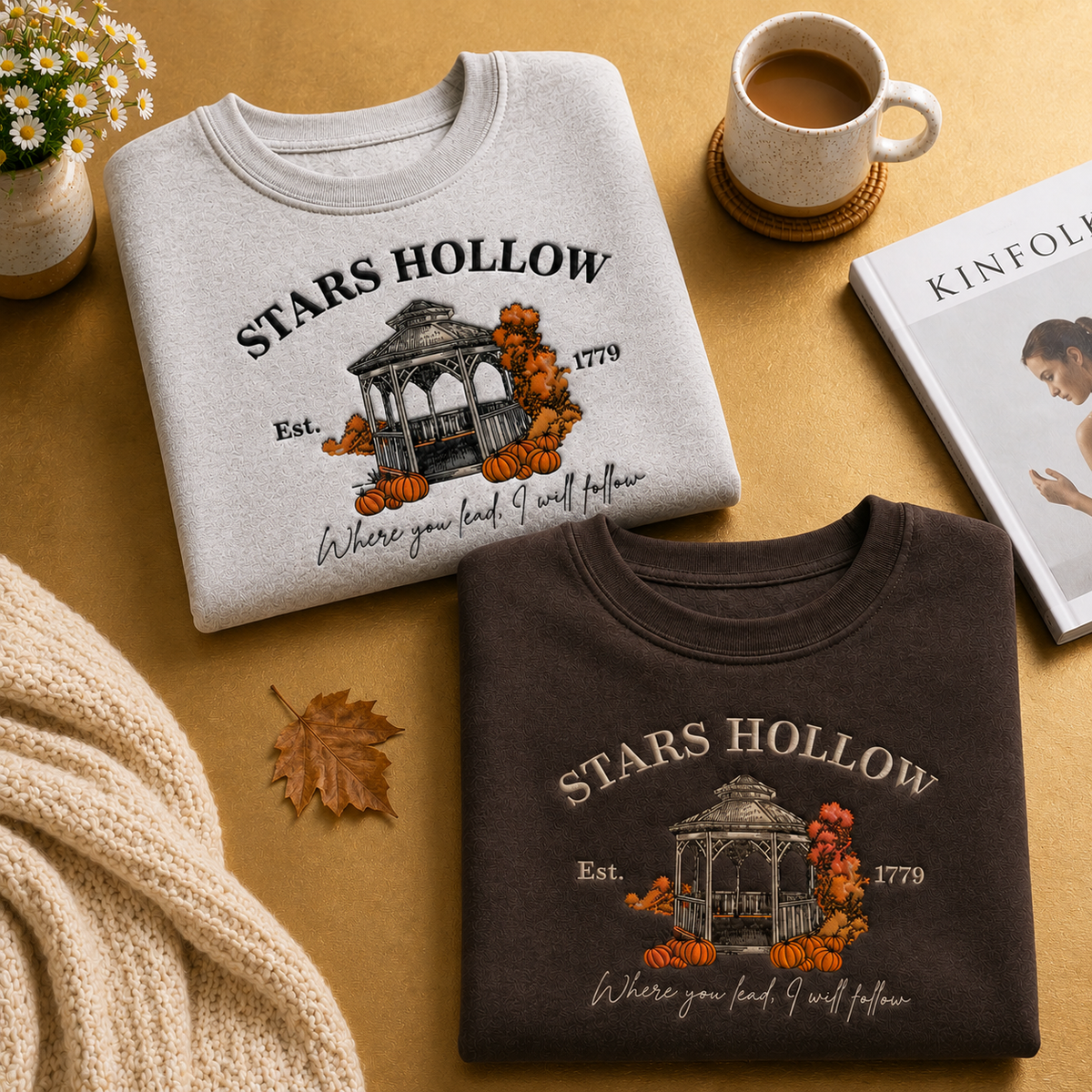 Two folded graphic T-shirts displayed on a warm tan surface with autumn props. The top shirt is light heather gray and the bottom shirt is dark brown, both featuring a vintage-style 'STARS HOLLOW' band graphic with a gazebo, pumpkins, and the text 'Est.