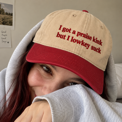 A person wearing a beige baseball cap with red embroidered text reading 'I got a praise kink but I lowkey suck' and a red visor, partially covered by a light gray hoodie in a bedroom setting.