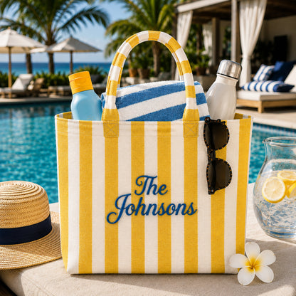 A yellow and white striped beach tote bag with blue embroidered text reading 'The Johnsons' sits on a poolside patio. The bag is styled with a blue-and-white striped towel, sunglasses, a water bottle, and other summer accessories, with a swimming pool, palm trees, and lounge chairs in the background.