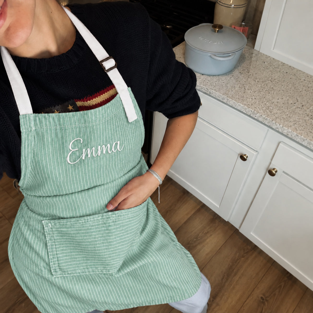 A person wearing a mint green and white striped kitchen apron with the name 'Emma' embroidered in white on the chest, standing in a kitchen next to a white Enamelware pot.