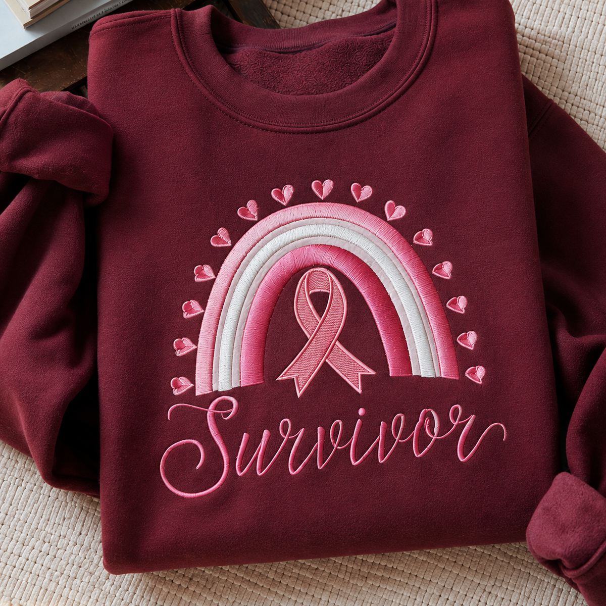 A folded burgundy sweatshirt with a pink and white rainbow, pink ribbon, heart pattern, and the word 'Survivor' embroidered on the front, displayed on a woven beige surface.