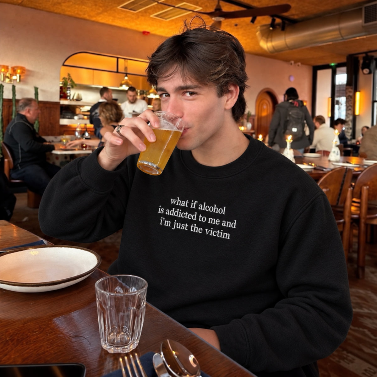 A man sitting at a restaurant table drinking from a glass, wearing a black crewneck sweatshirt with the white printed text 'what if alcohol is addicted to me and I'm just the victim'.